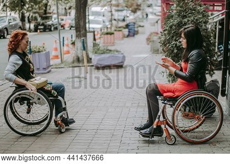 Two Girls In Wheelchairs Travel Through The Central Streets. Friends With Special Needs Communicate 