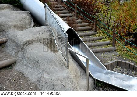 Stainless Steel Slide On The Field With Grassy Hills And Wooden Palisades. Large Glossy Tube For Sli