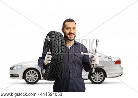 Auto mechanic worker in front of a silber car holding a tire on his shoulder and a lug wrench isolated on white background