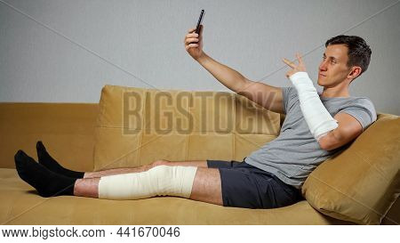 Young Brunet-haired Man In T-shirt With Damaged Leg And Broken Forearm In Gypsum Takes Selfies And P