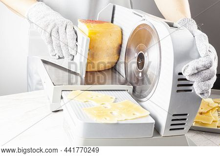 A Chef Is Slicing A Large Chunk Of Swiss Cheese Using An Electrical Deli Slicer. She Wears Protectiv