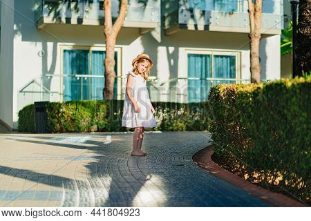 Little Girl In A White Dress And A Straw Hat Is Merrily Strolling Along The Path