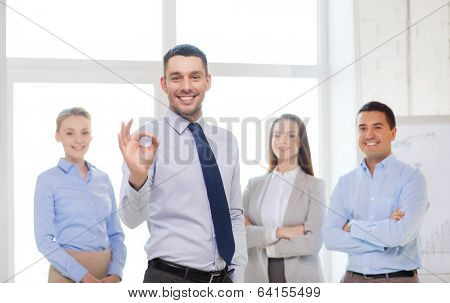 office, business, and teamwork concept - friendly young smiling businessman with team on back showing ok-sign