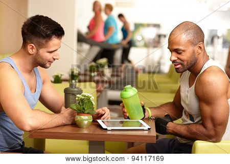 Athletic young men sitting at table in gym, taking a break, drinking refreshment, using tablet computer.