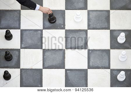 Close up of a player's hand, moving a black pawn forward during an outdoor chess game with large piiecs, seen from above