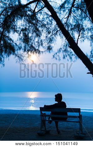 Solitude silhouette woman traveler sitting alone with her bag on retro bench at night on the beach.