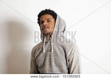 Close up of latino handsome guy wearing hood of his blank grey sweater looking weirdly on camera, isolated on white
