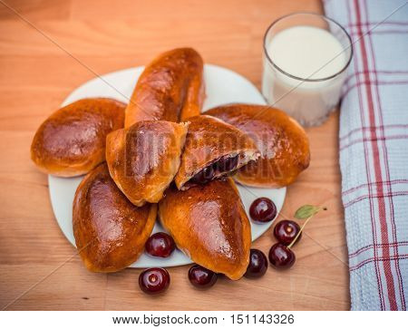 Oven fresh home made baked patties stuffed with cherry. Glass of milk. Delicious pastries on a baking sheet surrounded by cherry napkin 