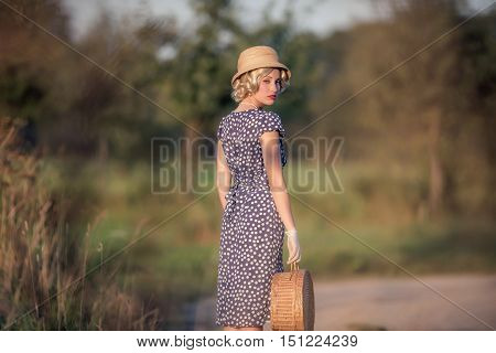 Retro 1930S Summer Fashion Woman With Blue Dress And Straw Hat Standing With Handbag In Rural Landsc
