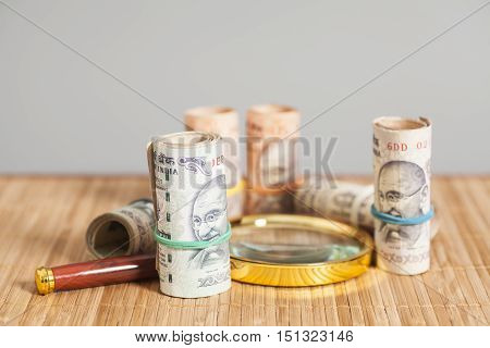 Rolls of Indian Currency Rupee Notes with magnifying glass on wooden background