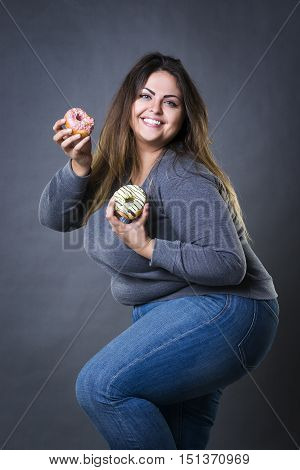 Happy beautiful young caucasian plus size model in blue jeans posing with donuts on a gray studio background fast food and unhealthy nutrition concept
