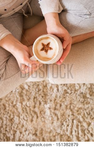 Girl with coffee mug sitting on the sofa indoors. Woman drinking a cup of coffee or tea sitting cozy at home. Relax and rest. Capuccino with cinnamon star. top view