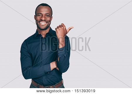 Look over there! Handsome young African man pointing away and smiling to you while standing against grey background