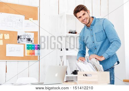 Smiling young man serching for documents in stack of folders on the office desk