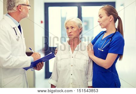 medicine, age, health care and people concept - male doktor with clipboard, young nurse and senior woman patient talking at hospital corridor