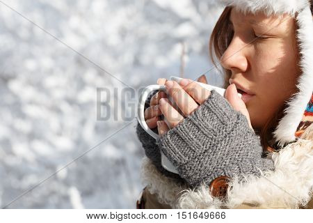 Beautiful young woman in gray fingerless knitted mittens cap with ear flaps and coat with white fur drinking tea or coffee holding white cup in hands at the background of snowy forest.
