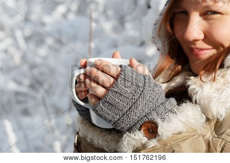 Beautiful young smiling woman in gray fingerless knitted mittens and coat with white fur holding in hands white cup of tea or coffee at the background of snowy forest.