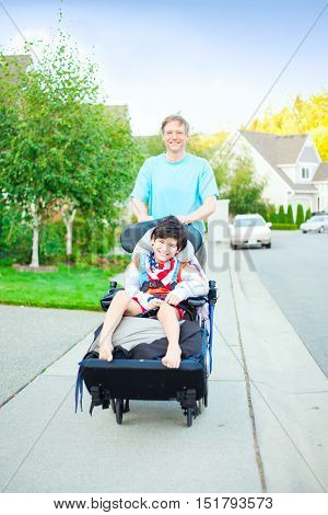 Caucasian father pushing disabled biracial ten year old son in wheelchair outdoors along sidewalk. Child has cerebral palsy.