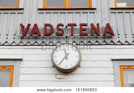 Close-up of public clockand the city name Vadstena on a gray railway station building.