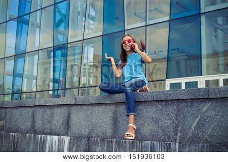 Excited and positive girl in lovely casual outfit has rather lively conversation on her smart phone while sitting on the concrete bench against the business center in the downtown 