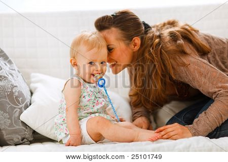 Adorable Baby mit Schnuller und junge Mutter spielen auf Couch