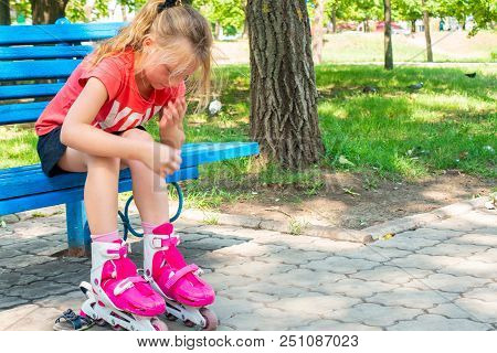 A Blond Girl Dresses Red Rollers, Sitting In A Park On A Blue Bench.