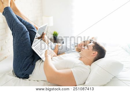 Mid Adult Man Using Laptop While Lying By Young Woman On Bed In Home