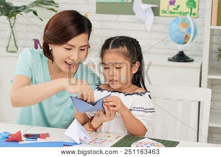 Cheerful Asian Teacher Woman And Little Girl Student Making Origami Ship In Art Class