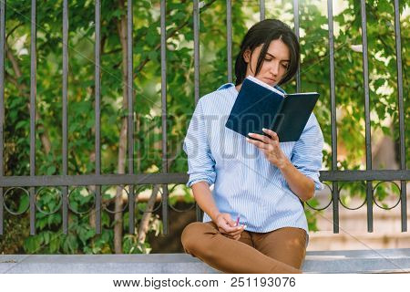 Serious Female Model Focused On Reading, Sits At Wall In The Park. Concetrated Student Prepares For 