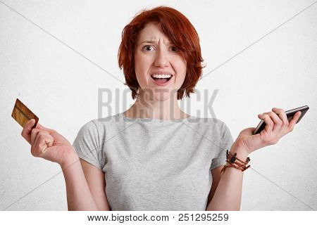 Indignant Ginger Woman Has Puzzled Expression, Holds In One Hand Smart Phone, In Other Credit Card, 