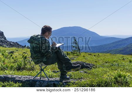 Traveler Sitting On A Camp Chair On A Mountain Plateau, Thinking And Writing Or Draws Something In A
