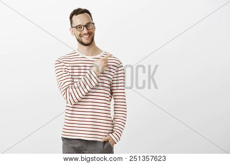 Portrat Of Proud Confident Aduly Guy In Glasses, Holding Hand In Pocket And Pointing At Upper Right 