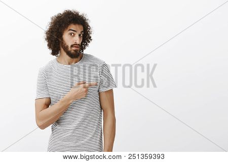 Indor Shot Of Unaware Doubtful Attractive Hispanic Guy With Beard And Afro Hairstyle, Pointing Right