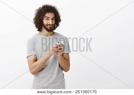 Playful Thougtful Handsome Guy With Curly Hair, Holding Smartphone, Smirking And Looking At Camera W