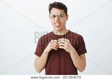 Studio Shot Of Funny Digusted Young Man In Round Glasses And Red T-shirt, Touching Fingers Nervously