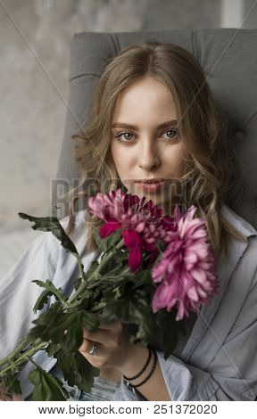 Portrait Of Blonde Beautiful Model With Curly Hair Sitting On The Grey Sofa. Girl Holding Pink Flowe