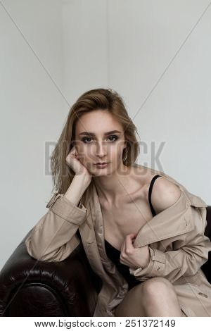 Beautiful Blonde Model Sitting In Brown Chair In Beige Raincoat. Girl Looking In Camera