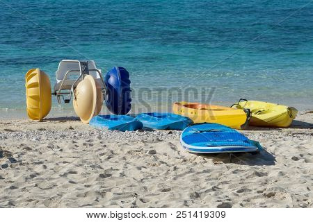 Kayaks, Paddle Boards, Surfboards, Paddle Boats, And Beach Toys In The Sand With Blue Water.