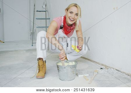 Happy young blond woman in white overalls and work boots sitting on hunkers with small yellow bottle mixing pastel wall paint in bucket at plastered wall and looking at camera.