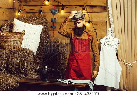 Handsome Man Tailor Or Dressmaker With Beard And Moustache In Apron Puts Hat On Head Near Vintage Se