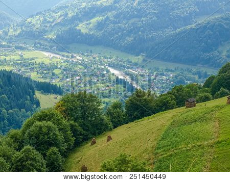 View Of The Mountain Village In Valley From A Mountain Slope With A Hayfields And Forest On The Fore