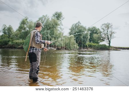 Adult Stands In Shallow And Holding Fly Rod In Hands. He Is Fishing. Guy Is Holding Fly Rod In One H