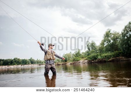 A Picture Of Man Holding Up Fly Rod In Right Hand. He Is Standing In Shallow Water And Looking Strai