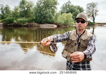 A Picture Of Cool Guy Stands In Water And Fishing. He Holds Fly-fishing With Reel Under It In One Ha