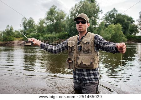 A Picture Of Brutal And Calm Guy Standing In Water And Looking Forward. He Has Fly Rod In One Hand A