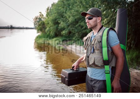 Serious Fisherman Is Standing In Shallow And Looking Straight Forward. He Holds Plastic Black Box In