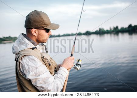 A Picture Of Man In Cap, Sunglasses And Vest Standing In Shallow And Looking At Fly Rod. He Is Using