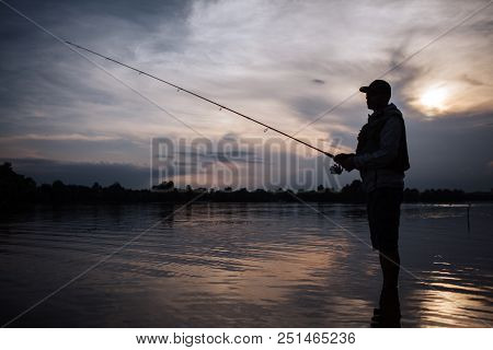 It Is Evening Outside. Fisherman Stands In Shallow And Fishing. He Holds Fly Rod In Hands. There Is 