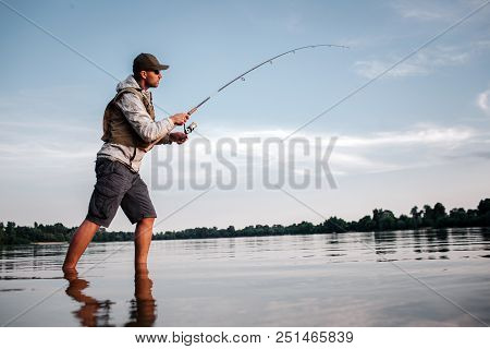 Active Man Is Standing In Shallow And Fishing. He Holds Fly Rod In Hands. Man Is Twisting Around Ree
