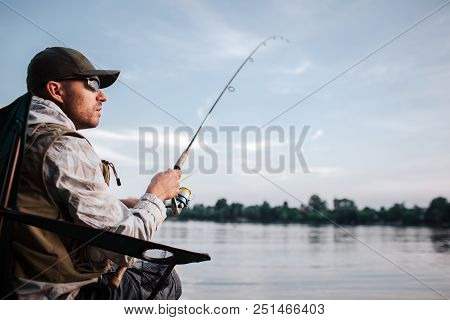 Thoughtful Guy Is Sitting At The Edge Of Water And Looking To The Right. He Holds Fly Rod In Hands. 